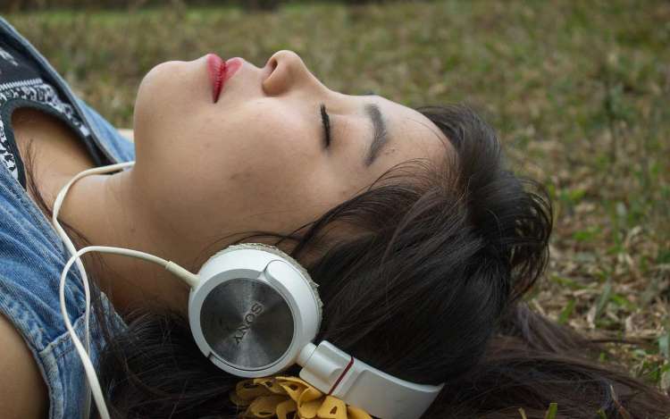 A woman wearing large headphones lays in the grass with her eyes closed, focusing on the music.
