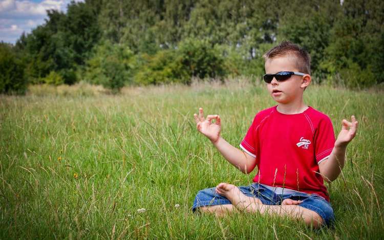 A young boy sits in the grass with sunglasses on, with his hands in a meditation mudra (hand gesture).