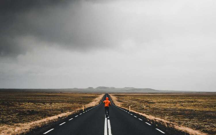 A person in an orange raincoat walking down the center line of a highway with storm clouds on the horizon.