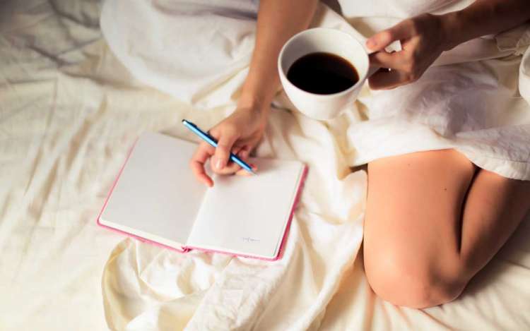 A person with a cup of coffee in one hand journals in bed.