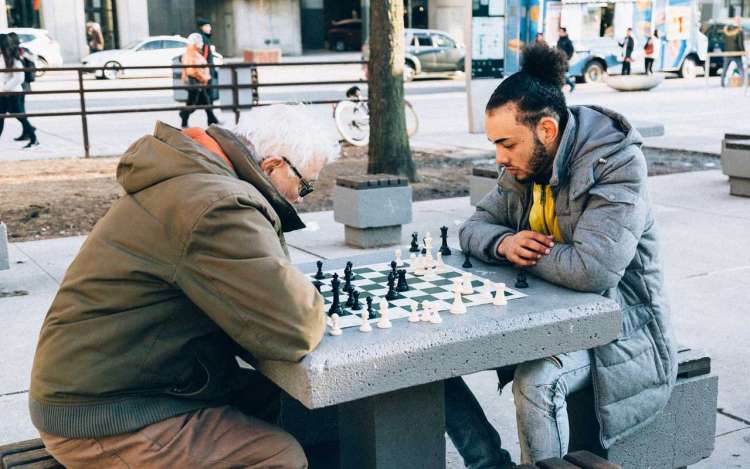 An older man plays chess in the park with a younger man. Chess is one type of cognitive activities for adults that may be beneficial as you age.