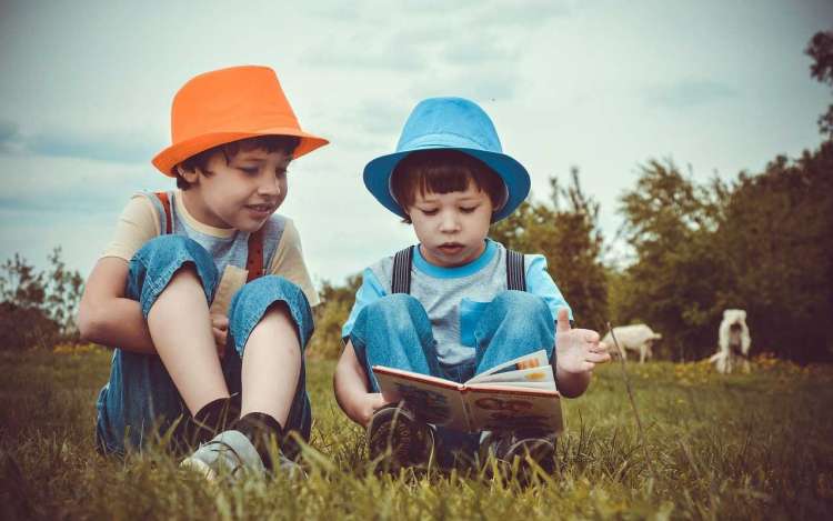 Two young brothers read out loud from a book while sitting in the grass.