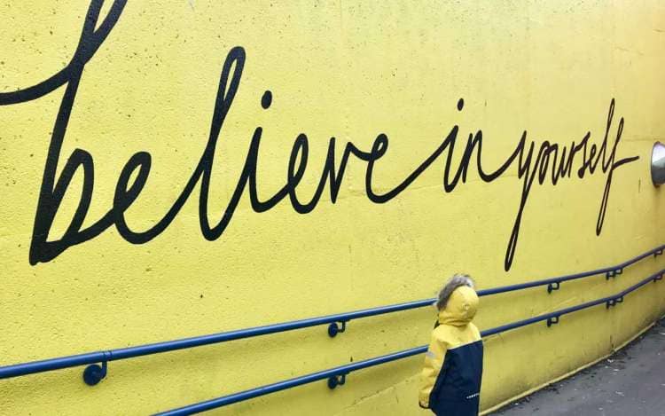 A "believe in yourself" mural painted on a bright yellow wall, with a small child looking at the wall.