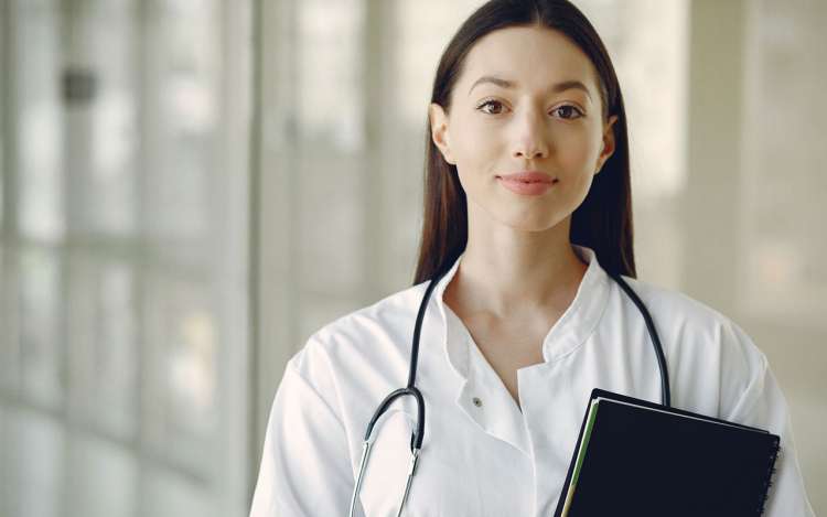 A doctor stands in a hallway with her stethoscope around her neck and a notebook in her hand.