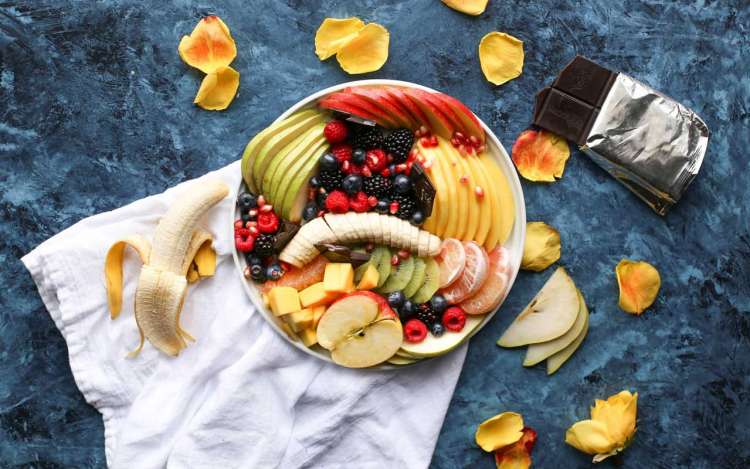 A plate full of different kinds of fruits like bananas, apples, berries, and citrus sits next to a bar of dark chocolate. Healthy foods can boost memory.