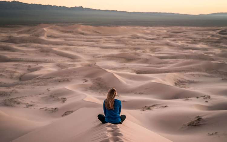 A woman sits and meditates on top of a sand dune in the desert. This could be a good setting for a guided visualization.