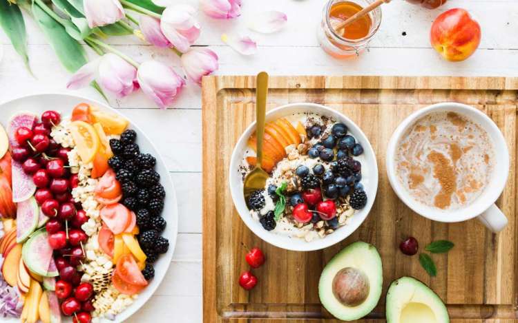 Plates of fruits and vegetables and a bowl of oatmeal with fruit sit on a table with tulips and a wooden cutting board.