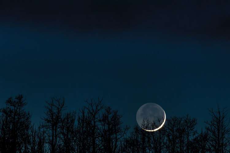 A sliver of a crescent moon hangs low on the horizon at dark, just visible over the tree line.