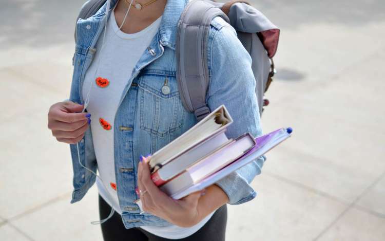 A woman wearing a denim jacket holds a stack of books as she listens to headphones.