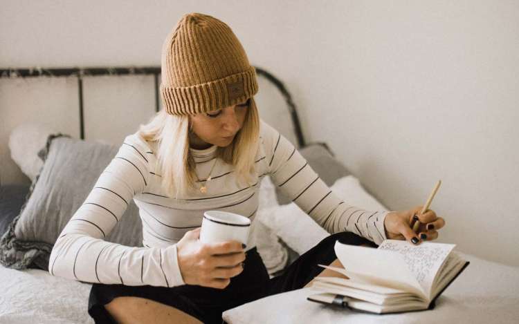 A woman wearing comfortable clothing and a beanie hat studies on her bed.