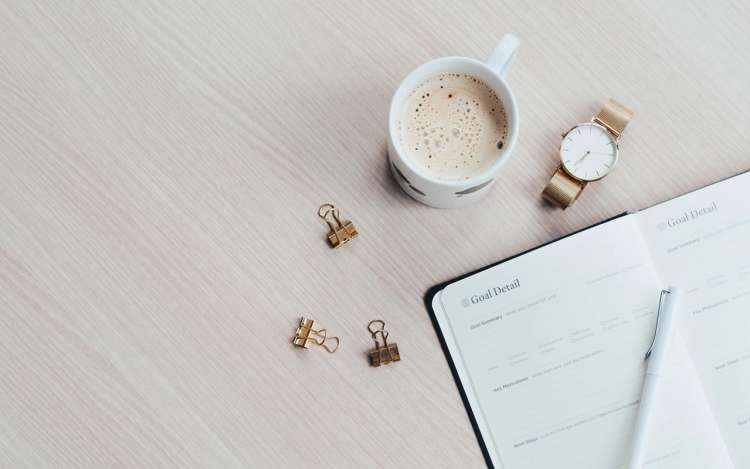 A latte and a watch sit on a desk next to a paper goal planner.