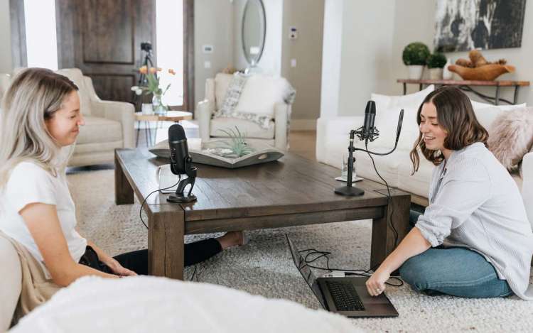 Two women sit in a living room and prepare to record a podcast episode.