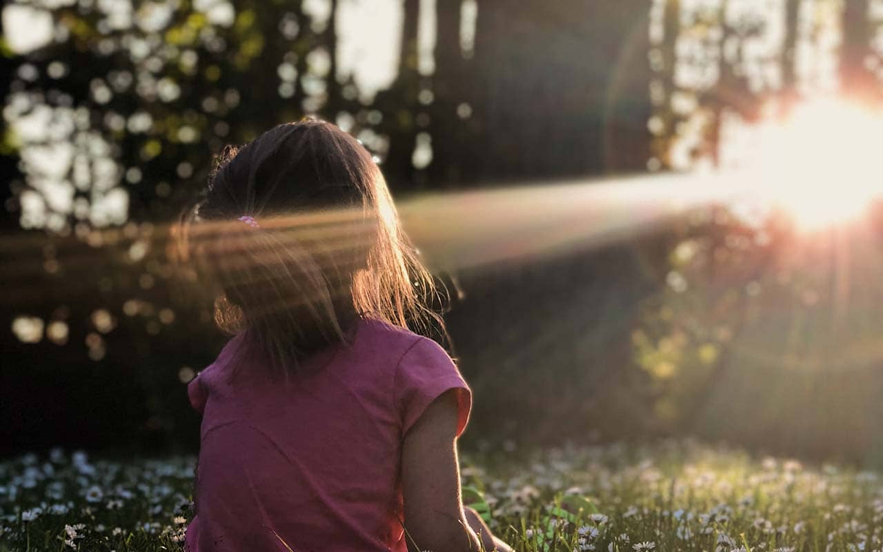 A young girl sits in a beam of light in nature. Choosing a natural environment to meditate in can help you focus.