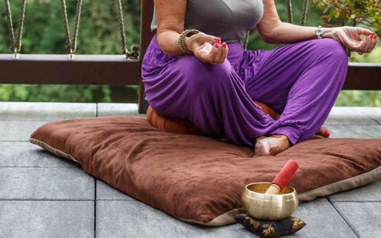 A woman in purple pants sits on a meditation cushion with her hands in a mudra, with a singing bowl next to her.