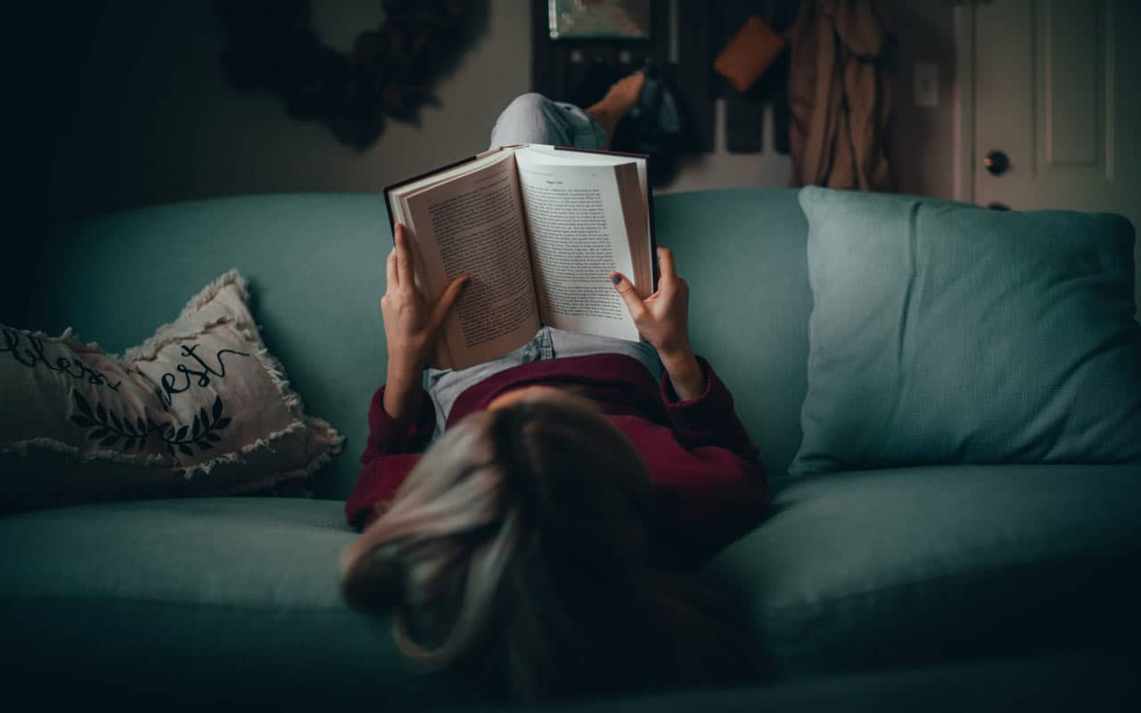 A person reads on a couch with their legs up over the back of the couch.