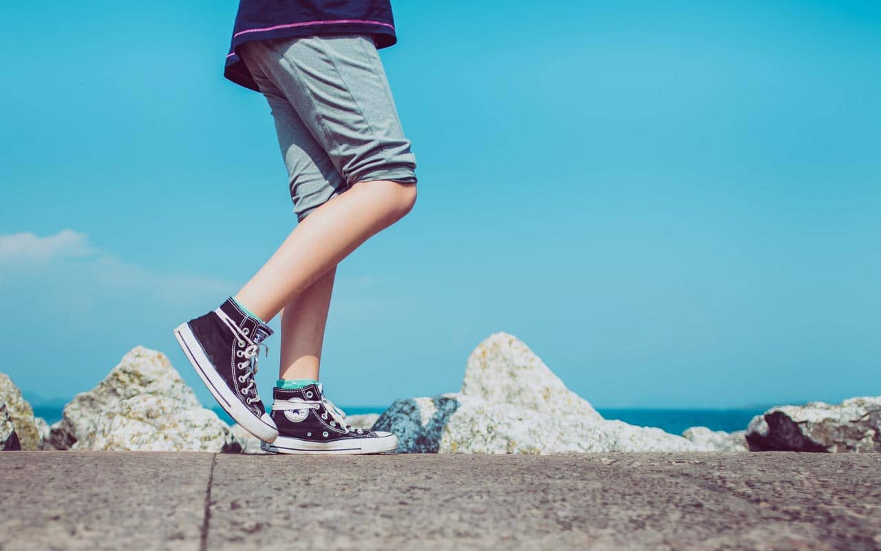 A person wearing Chuck Taylors walks along a concrete sidewalk, against the backdrop of a blue sky.