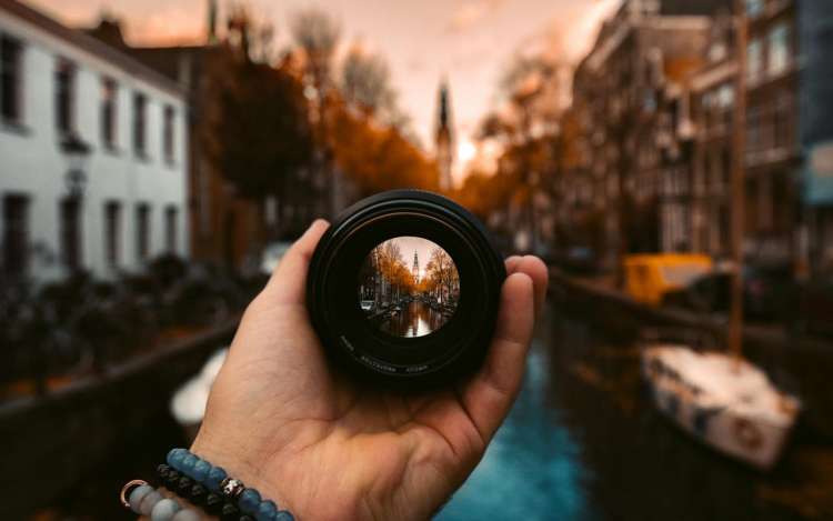 A person holds a camera lens. The focus shows a canal in Amsterdam, with the rest of the photo in soft focus.