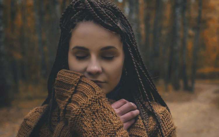 A woman with braids stands on a forest path in a chunky sweater with her eyes closed.
