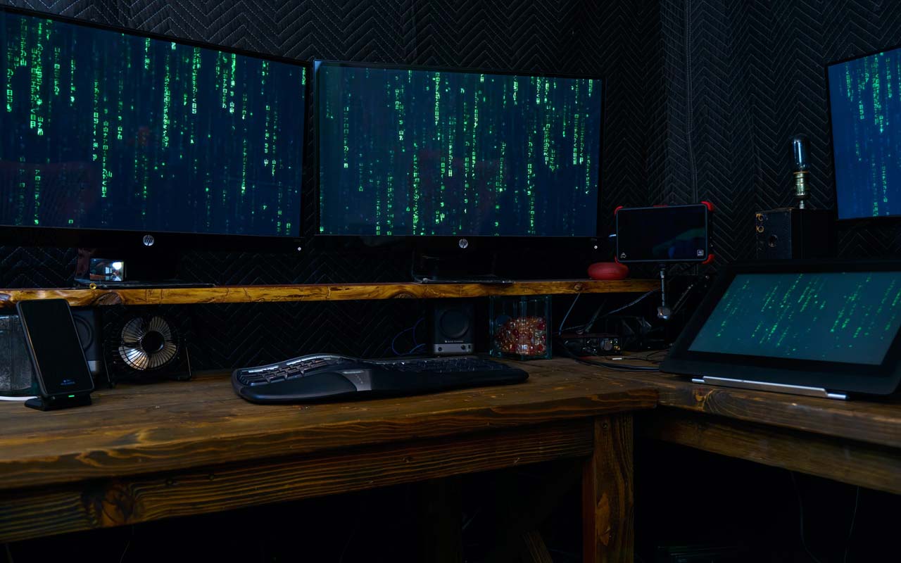 Multiple computer screens sit on a desk, running code that looks like the type used in the movie The Matrix.