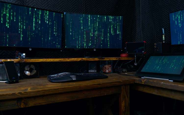 Multiple computer screens sit on a desk, running code that looks like the type used in the movie The Matrix.