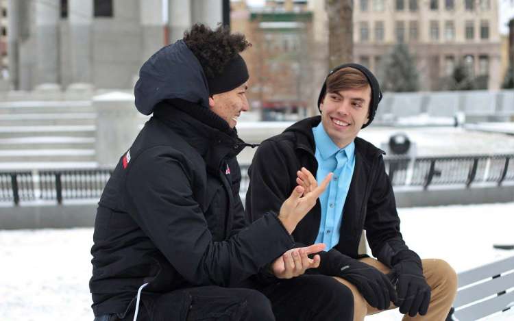 Two young men sit outside on a bench, conversing. One is smiling and listening while the other talks and uses hand gestures.