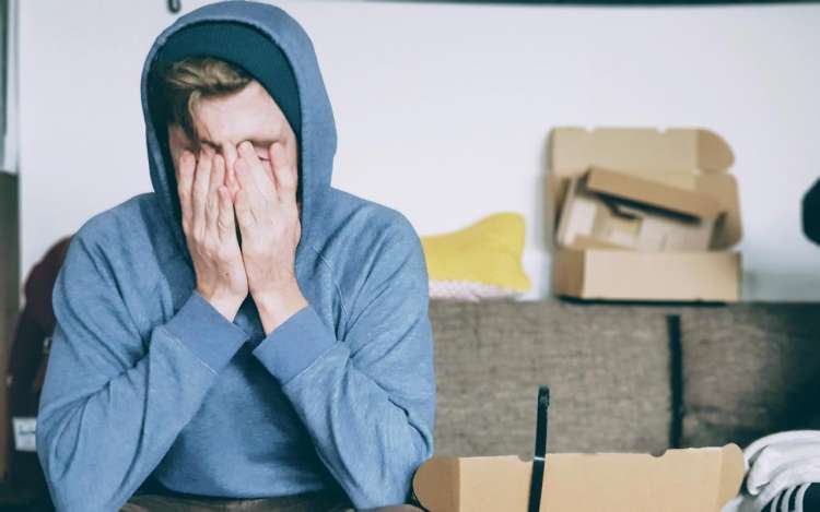A person wearing a blue hoodie sits on a couch with their hands over their face.