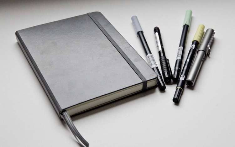 A black day planner sits on a table with a handful of pens. Writing out your daily plan in Spanish is a good way to practice your language learning.