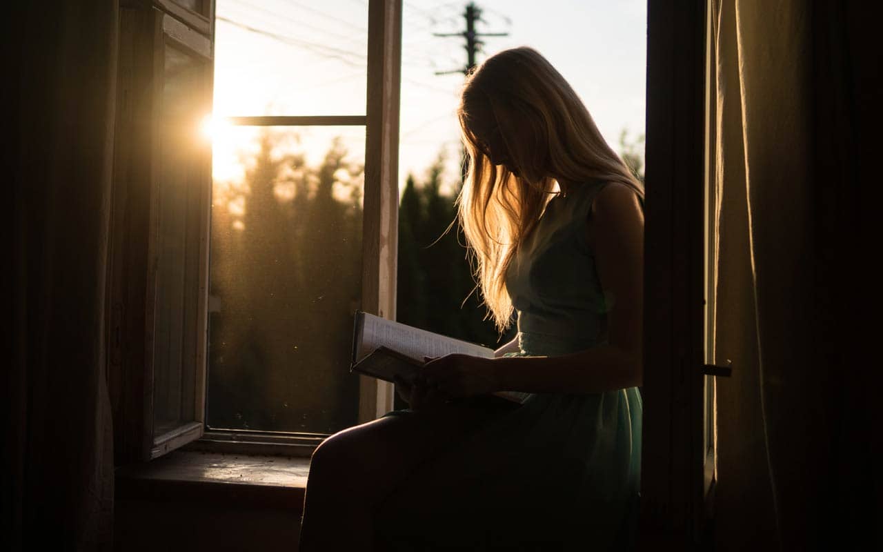 At sunset, a woman sits near an open window with a book.
