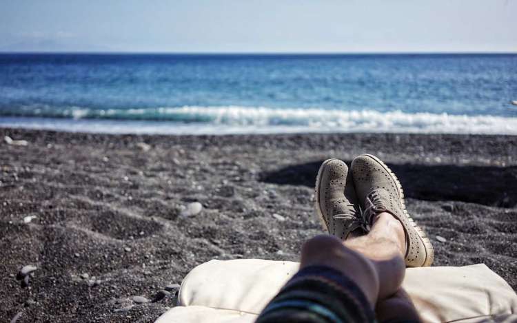 A person relaxes at a black sand beach with their shoes on and legs up.