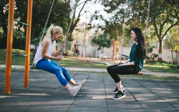 Two women sit on swings outside in a park. Speaking is one of the best ways to learn Spanish quickly.