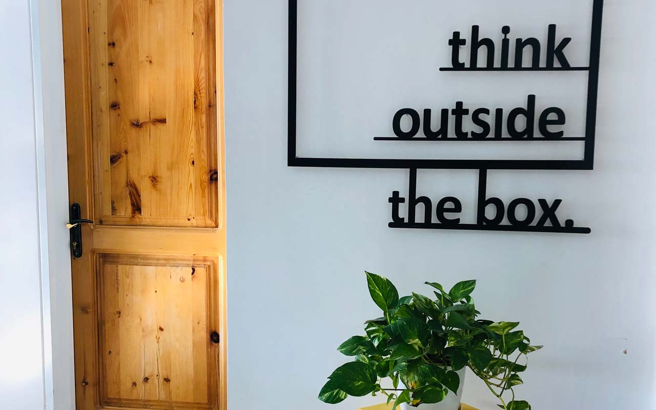 A sign with the text "think outside the box" against a white wall, with a green plant on a table underneath the sign.