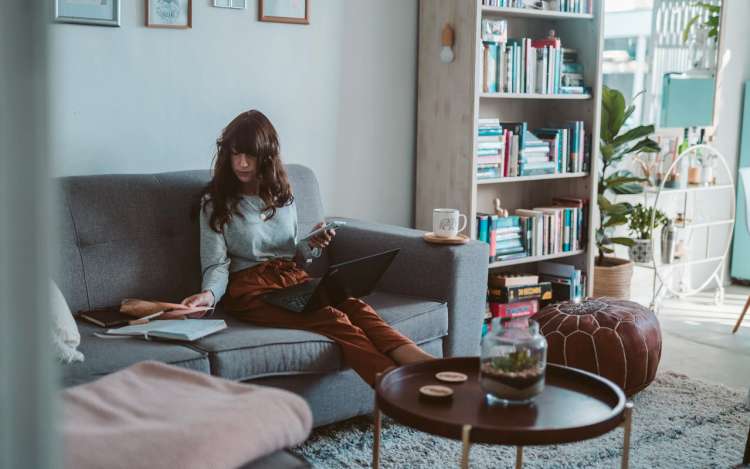 a woman sits on sofa with a book and a laptop